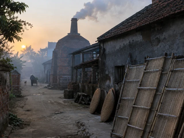 A craft-village lane at dawn, bamboo drying racks along the wall, a kiln chimney smoking, Red River Delta mist