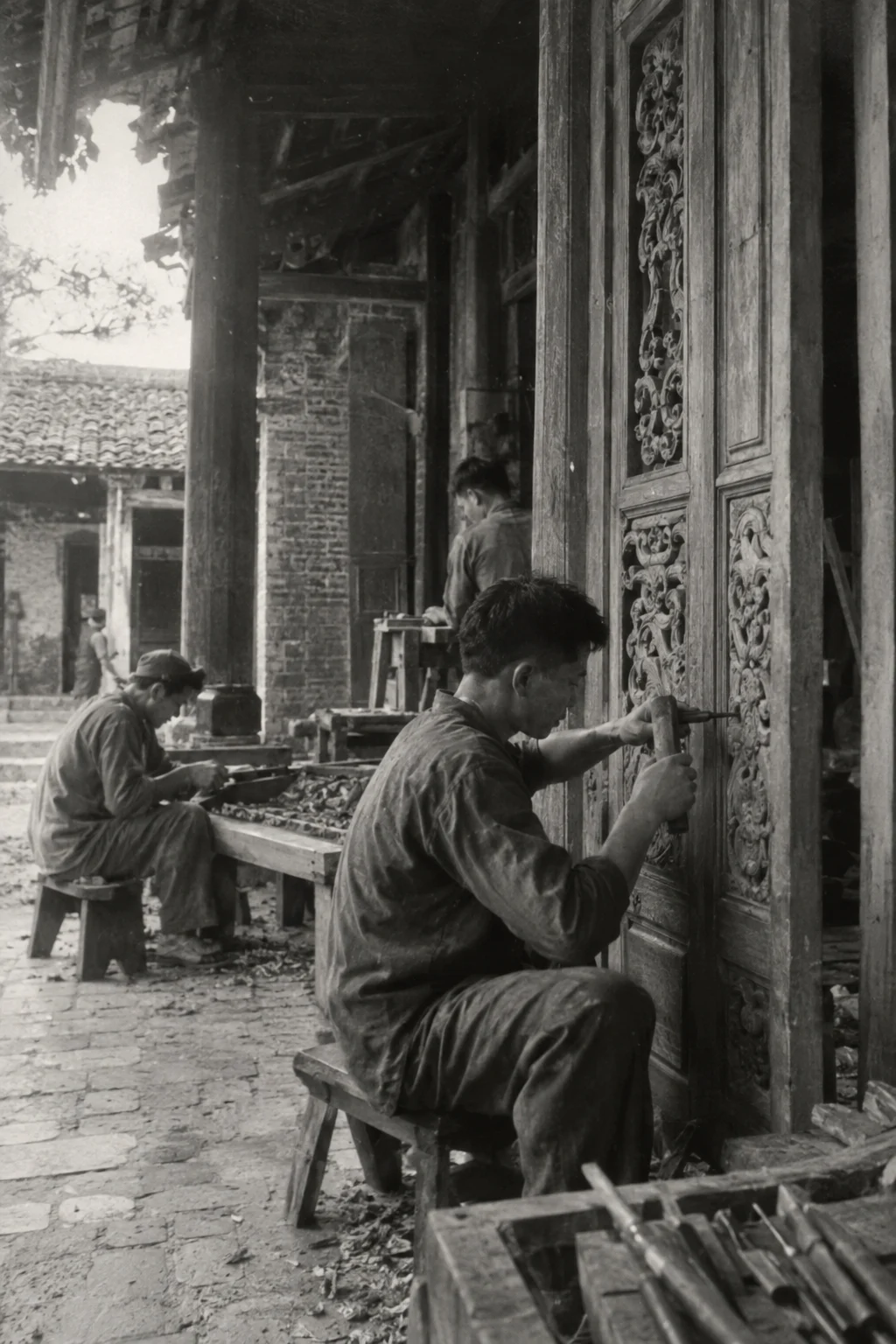 Archive black-and-white photograph from the mid-twentieth century, faded and grainy: carving a door in Đồng Kỵ, carved hardwood, villagers in simple work clothes, old brick and timber