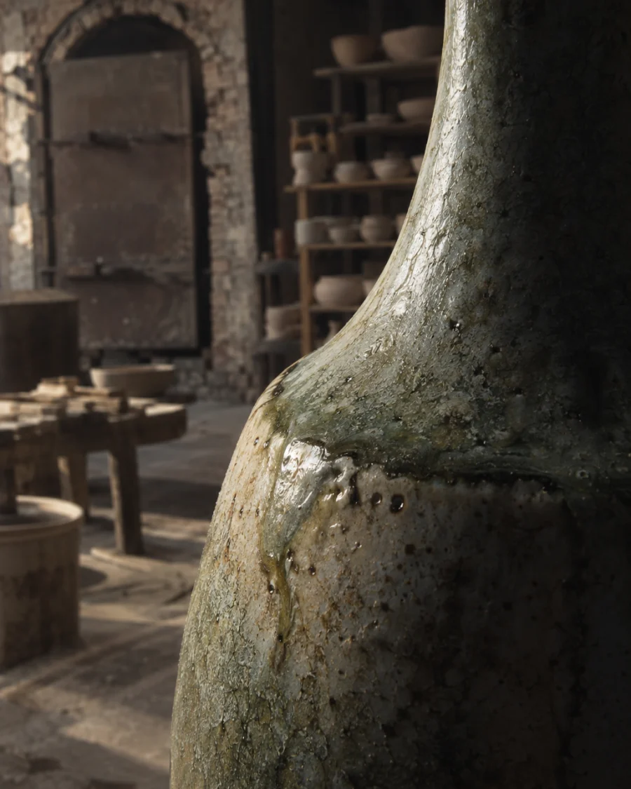 Bottle vase, ash glaze, close-up of the shoulder where the form turns, glaze pooling, in a Bát Tràng pottery workshop, brick kiln door and shelves of bisque ware behind, sage greens and warm greys