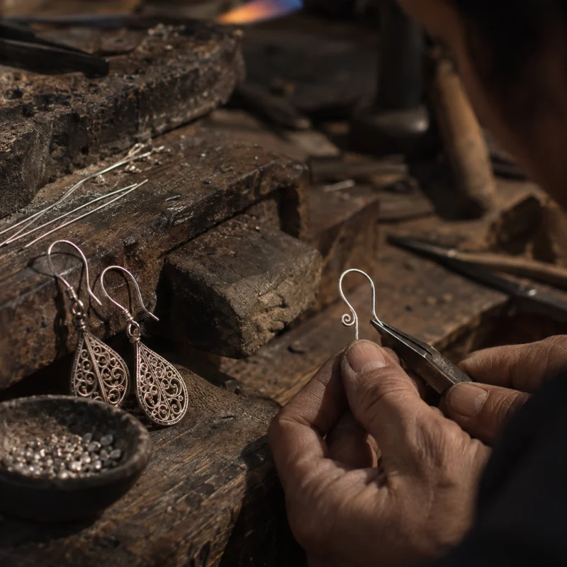 Filigree drop earrings, close-up of the hand-formed silver ear hooks, at a Định Công silversmith's bench, worn wood, fine silver wire and a small torch flame, warm grey and silver tones