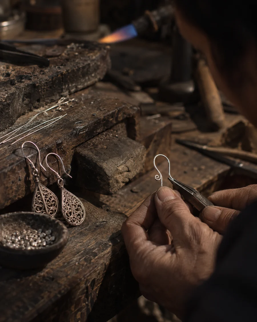 Filigree drop earrings, close-up of the hand-formed silver ear hooks, at a Định Công silversmith's bench, worn wood, fine silver wire and a small torch flame, warm grey and silver tones