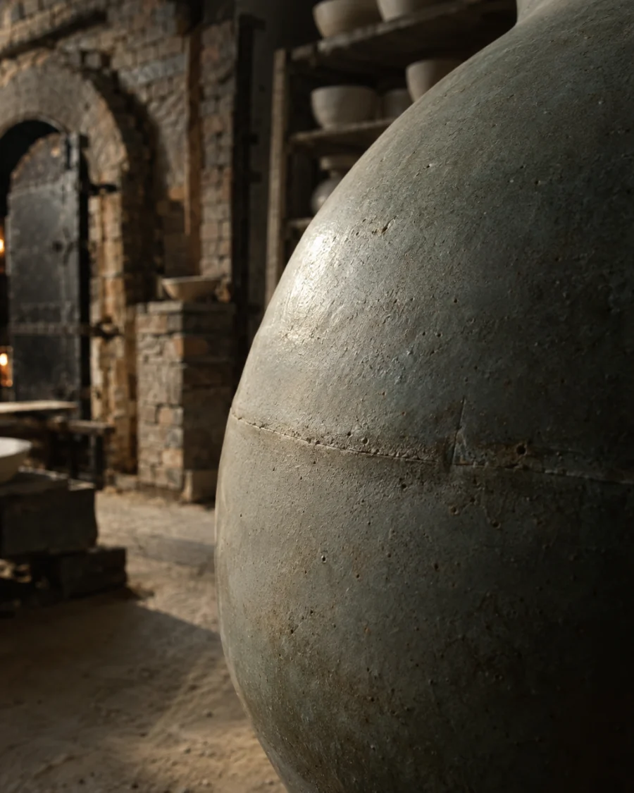 Moon jar, ash glaze, close-up of the faint seam where the two thrown halves meet, in a Bát Tràng pottery workshop, brick kiln door and shelves of bisque ware behind, sage greens and warm greys